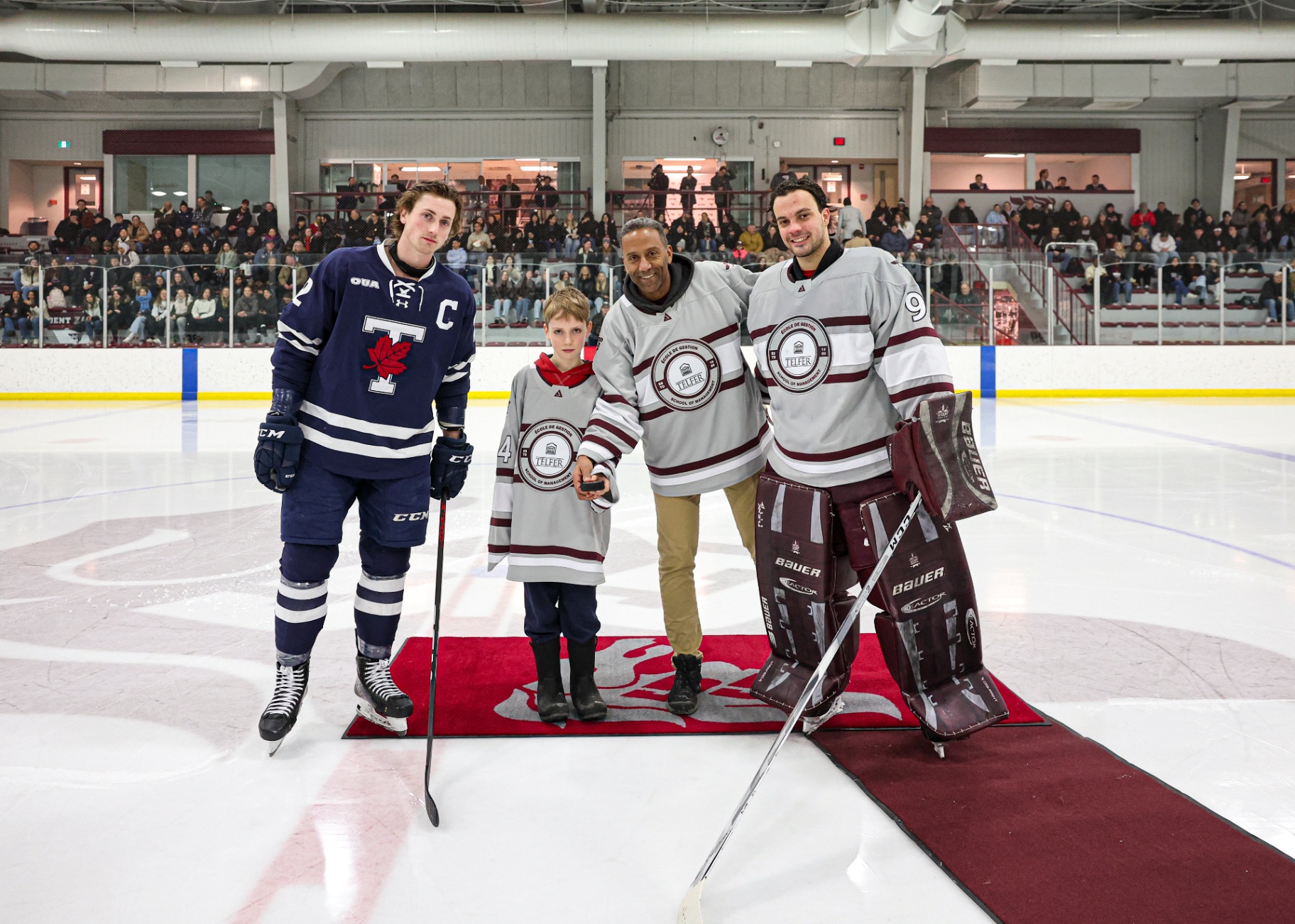 Franky Lapenna smiles and holds a hockey stick while standing on the ice beside the dean and a young boy holding a puck. On the left is a player from the University of Toronto.