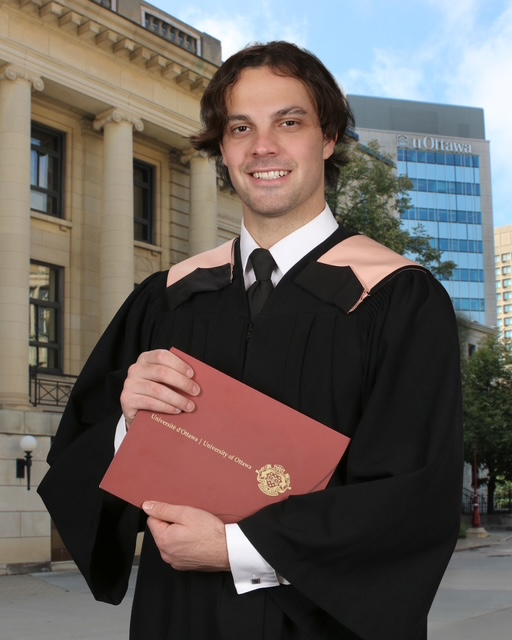 Franky Lapenna smiles in a graduation gown while holding his diploma, with the Desmarais Building and Tabaret Hall behind him.