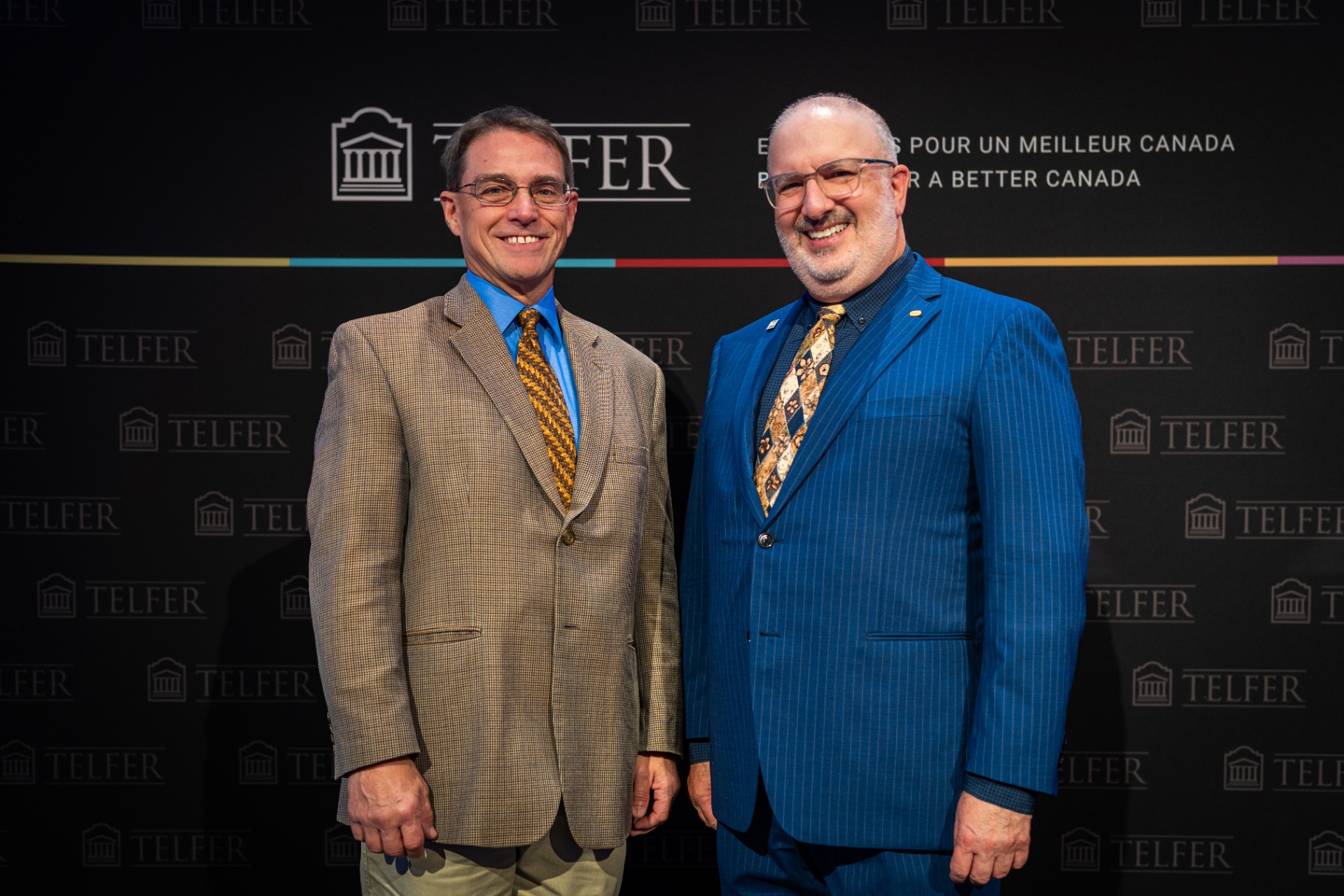 Jeffrey R. Smith (à gauche), portant un blazer marron à motifs, une chemise bleue et une cravate dorée à motifs, se tient aux côtés de Murray Kronick (à droite), qui porte un costume rayé bleu vif, une chemise sombre et une cravate à motifs, tous deux souriant devant un fond sombre avec la mention « TELFER »