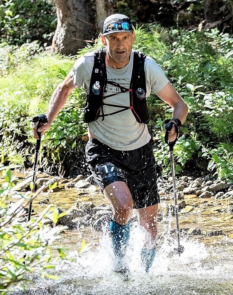 Myron Tetreault wearing a running vest and shorts races through a rocky stream with cross country skis.