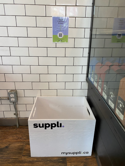 A drop-off bin with Suppli written on the front in front of a brick wall.
