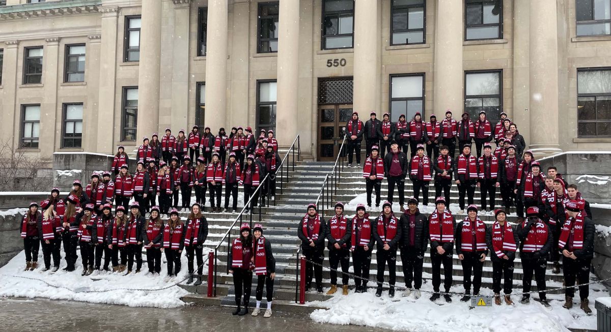 A large group of students wearing maroon scarves and all black clothing form the numbers 2 and 3 on the steps of Tabaret Hall.