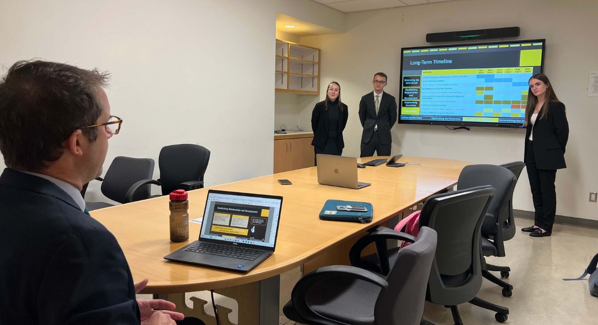 A professor at the end of a long table observes three students dressed in formal wear practising a PowerPoint presentation in a boardroom.