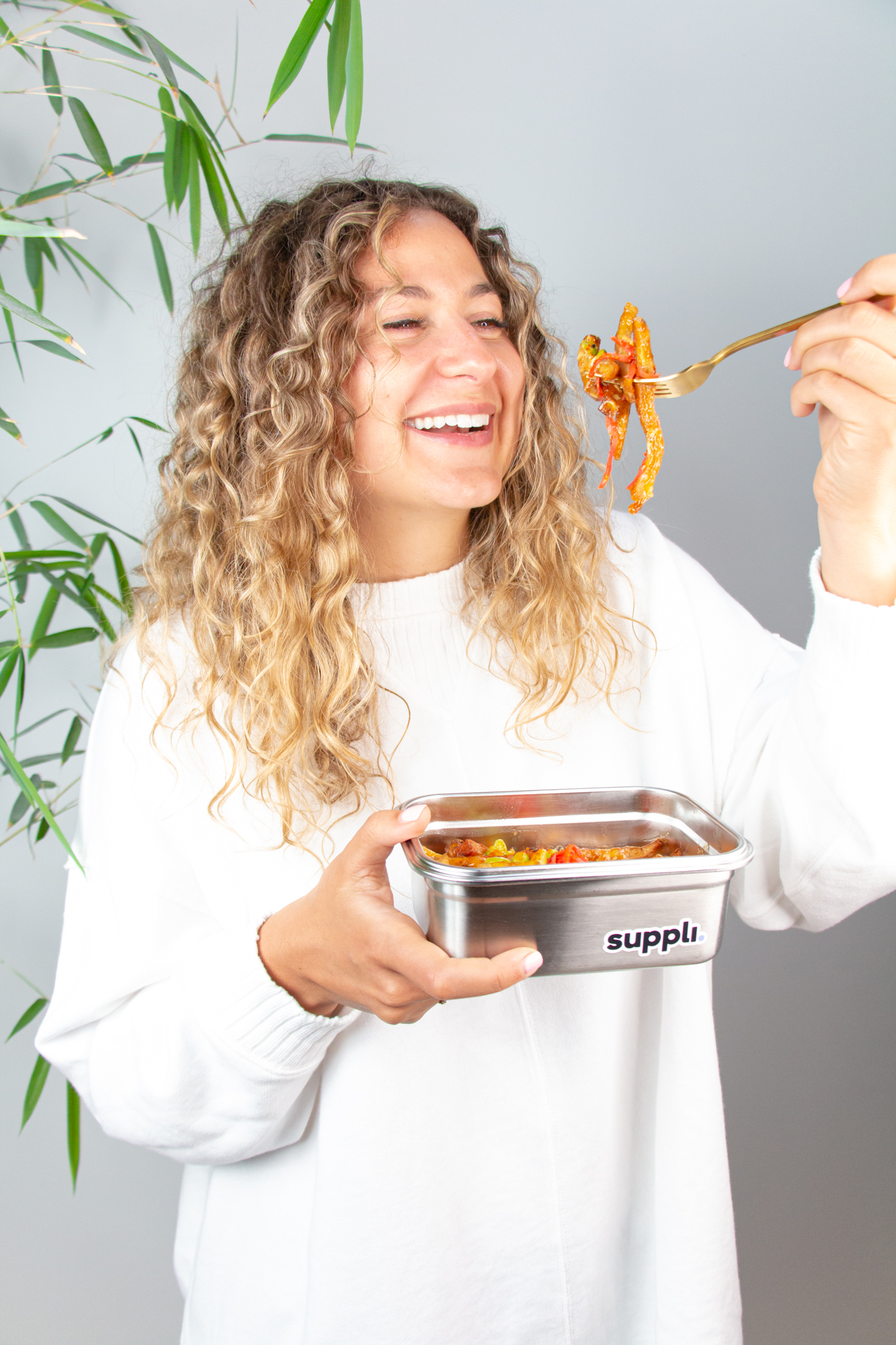 A smiling model wearing a sweater dangling food from a Suppli container on her fork.