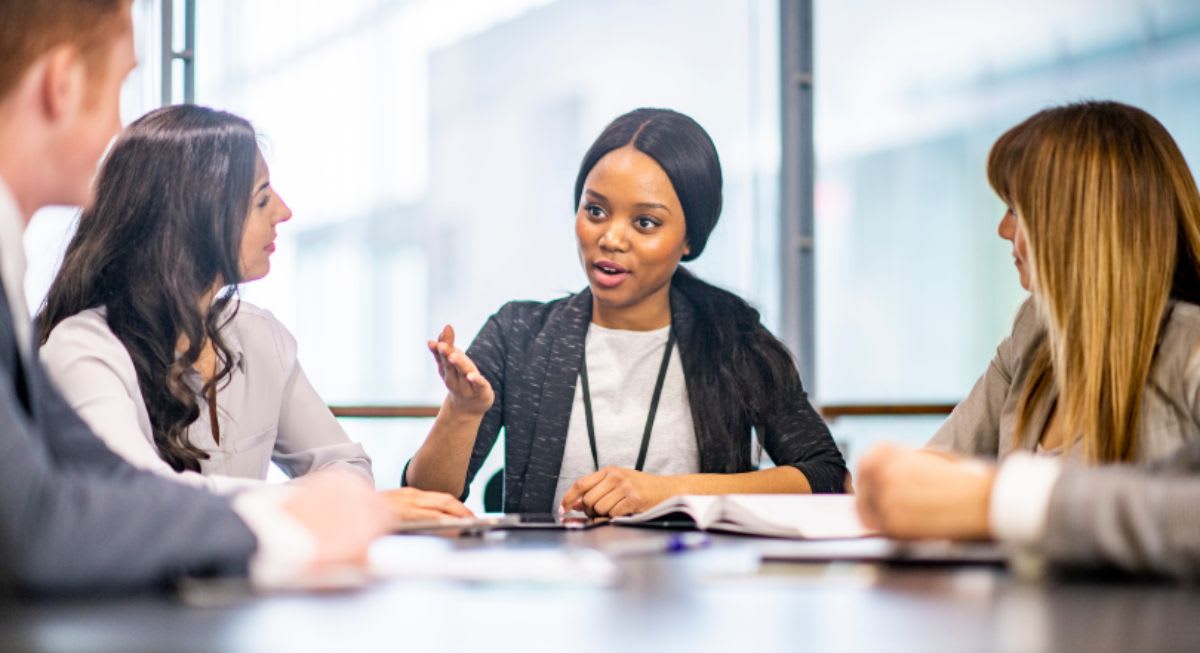 A woman sits at the head of a table in an office and addresses three colleagues sitting with her.