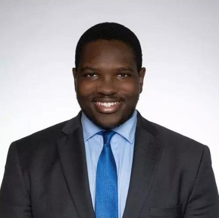 Calvin Diallo smiling wearing a black suit, light blue dress shirt and blue tie.