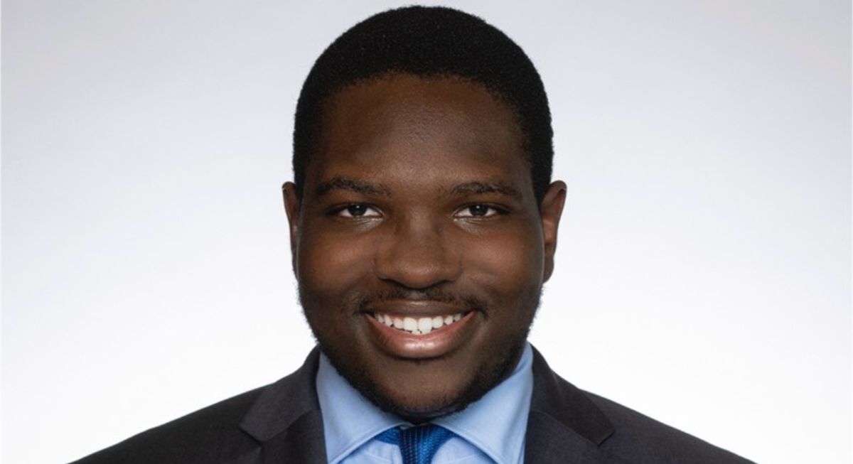 Calvin Diallo, wearing a dark grey suit, light blue shirt and blue tie, and smiling at the camera.