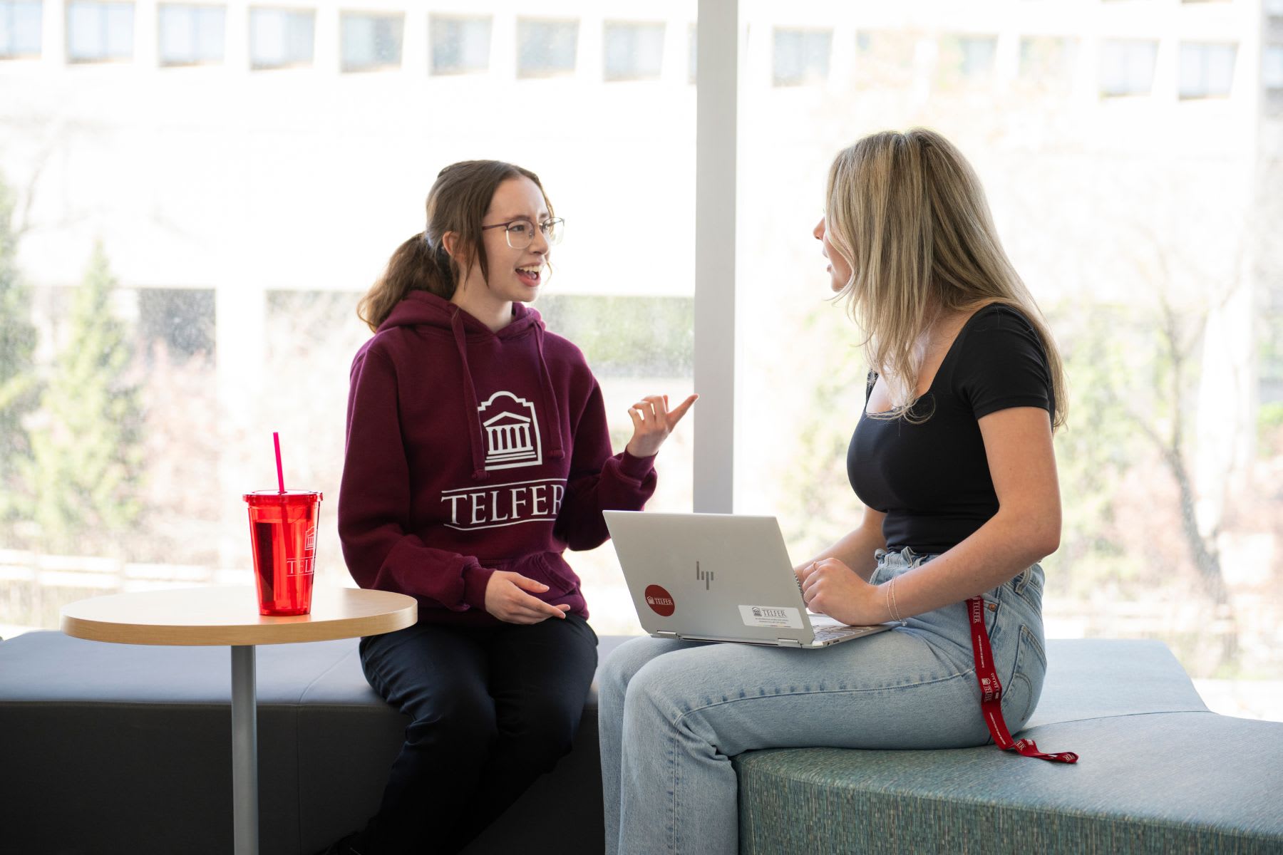 Deux étudiantes au B.Com. de l’École Telfer sont assises face à face dans le salon étudiant et se sourient en discutant. L’une des étudiantes porte un chandail à capuchon grenat arborant le logo de l’École Telfer. L’autre étudiante a un ordinateur portable sur les genoux.