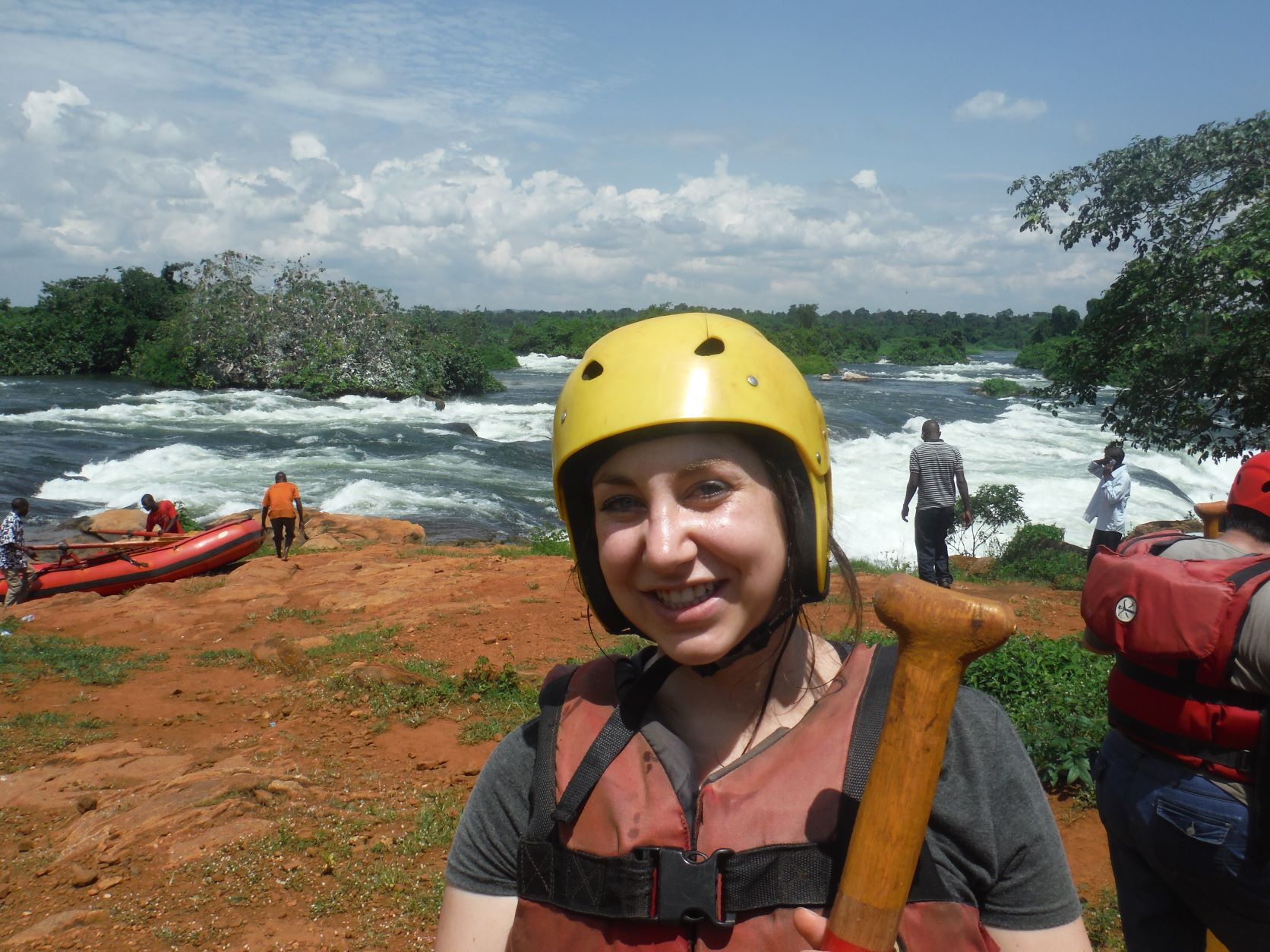 Katherine standing in front of a river in Uganda, wearing a yellow helmet and holding a paddle.