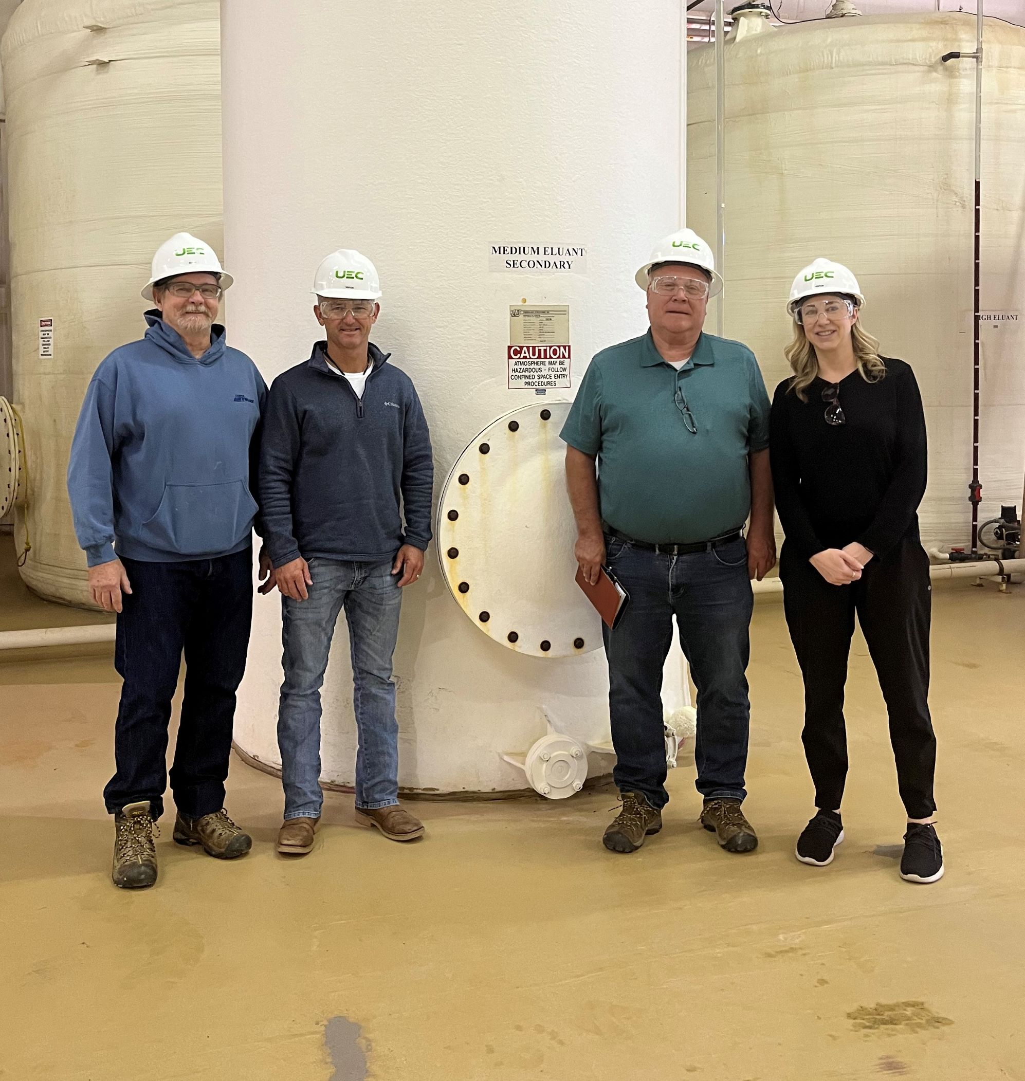 Katherine standing with three male colleagues, all in hardhats, in front of a tall barrel used to process uranium.