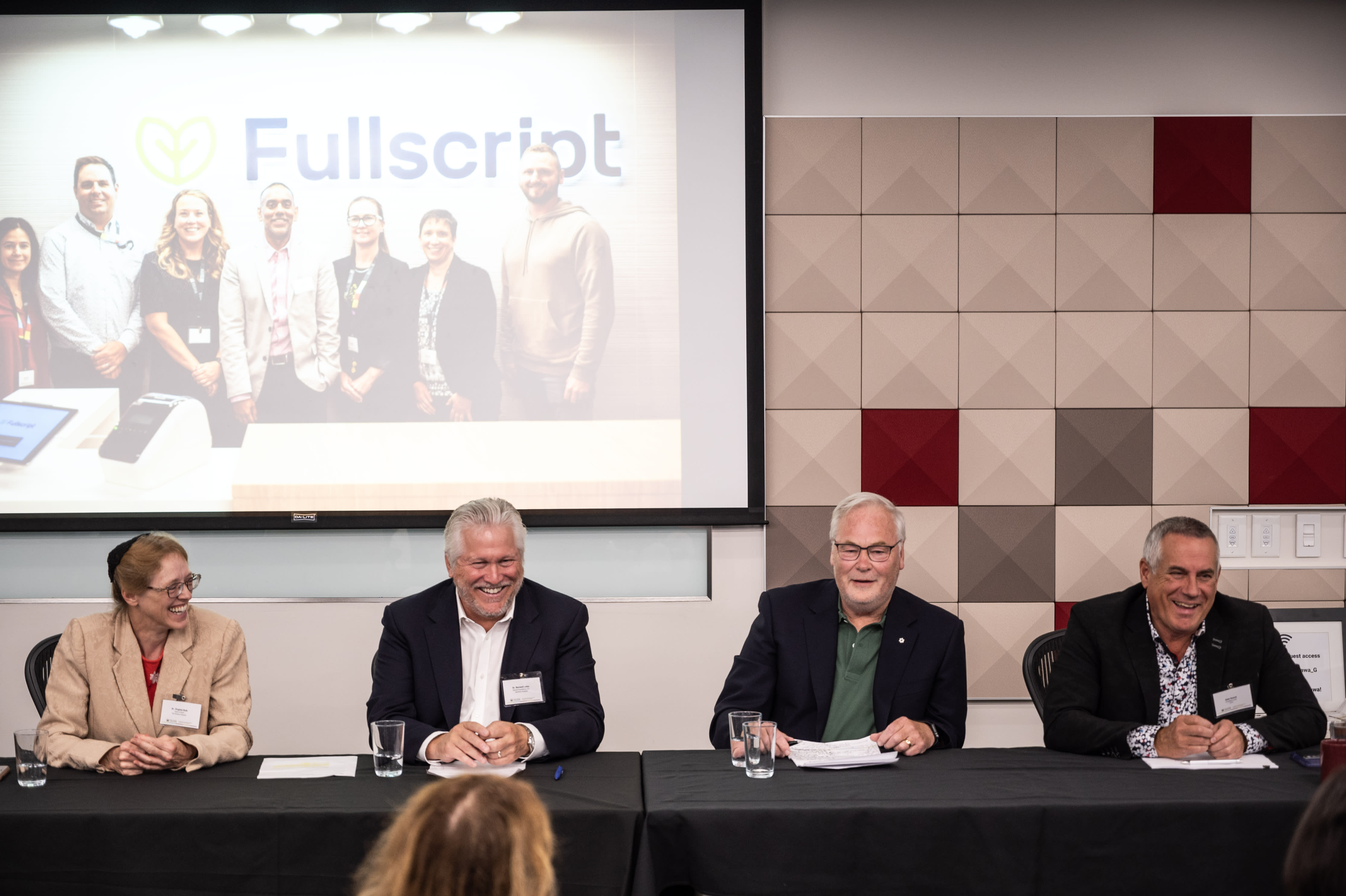 Four panelists in conversation and smiling while seated in a row at a table facing the seated attendees.