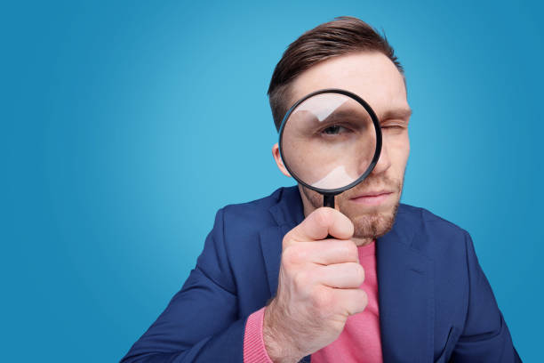 Man wearing dark blue suit and pink sweater holding a magnifying glass to his right eye.