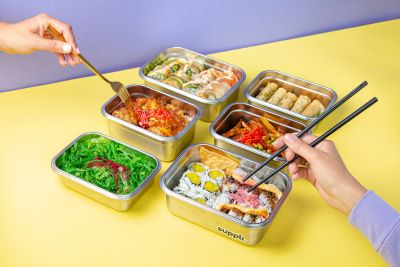 Suppli containers on a table, with the hands of two people eating out of them, using a fork and chopsticks, respectively. Containers are stainless steel and filled with different types of food.