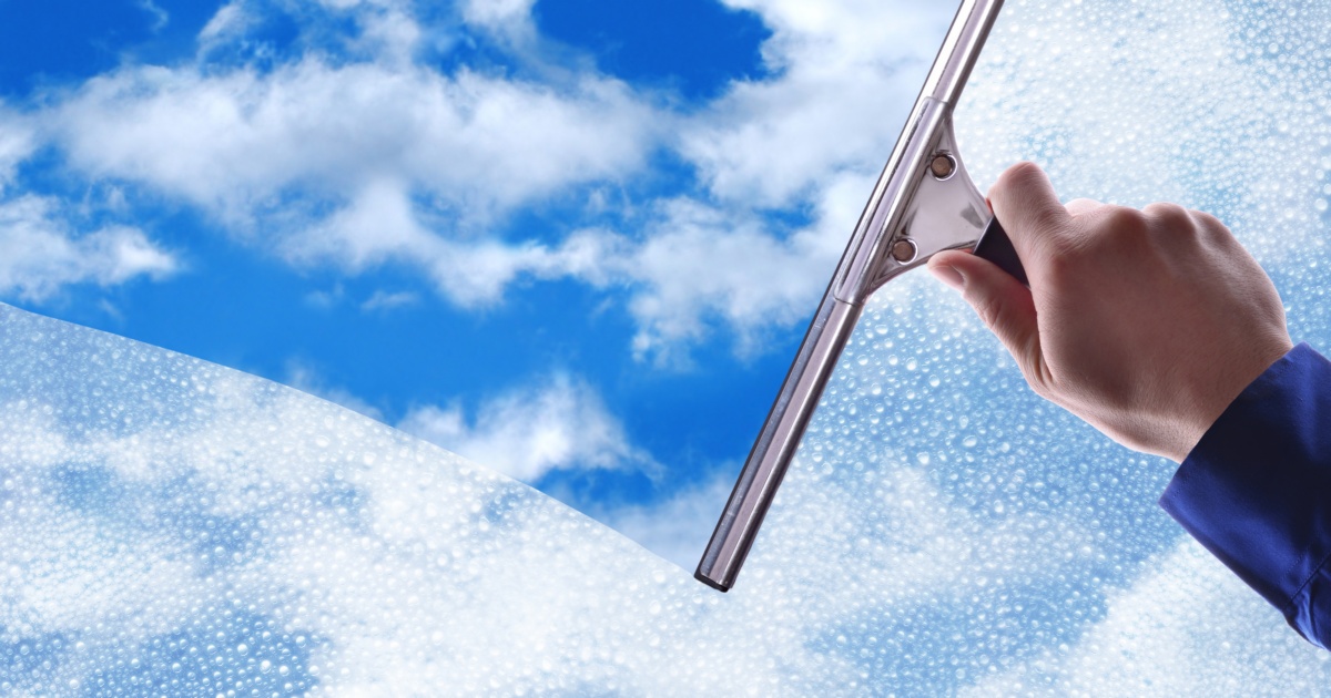 The hand of someone holding a wiper, cleaning a window showing a blue slightly clouded sky.