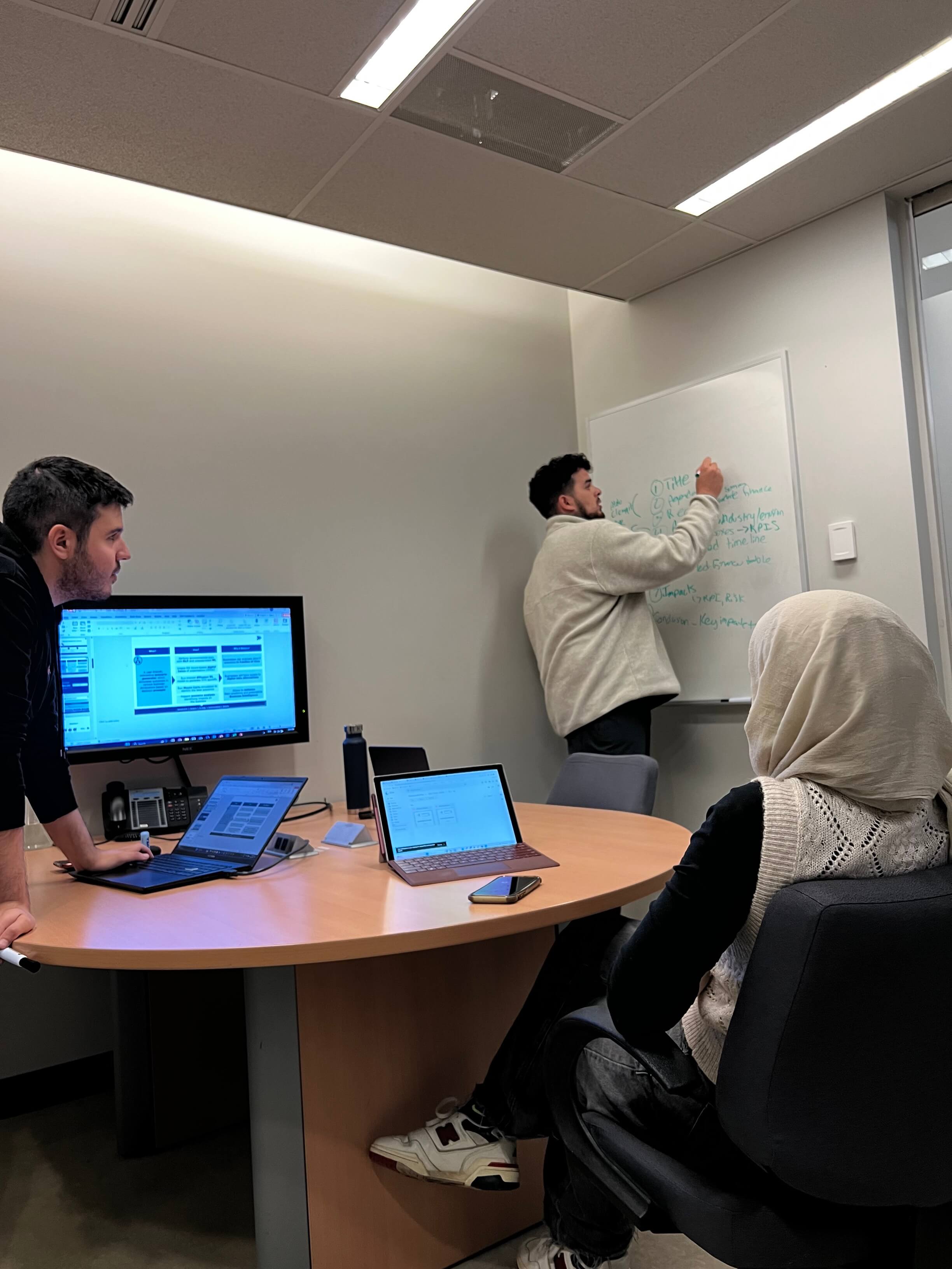 Three students in a boardroom prepare for a presentation. One student writes on a whiteboard. Two students watch.